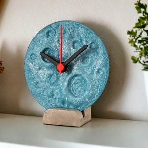 May include: A teal blue clock with a textured surface resembling the moon. The clock has black hands and a red second hand. The clock is on a concrete stand.