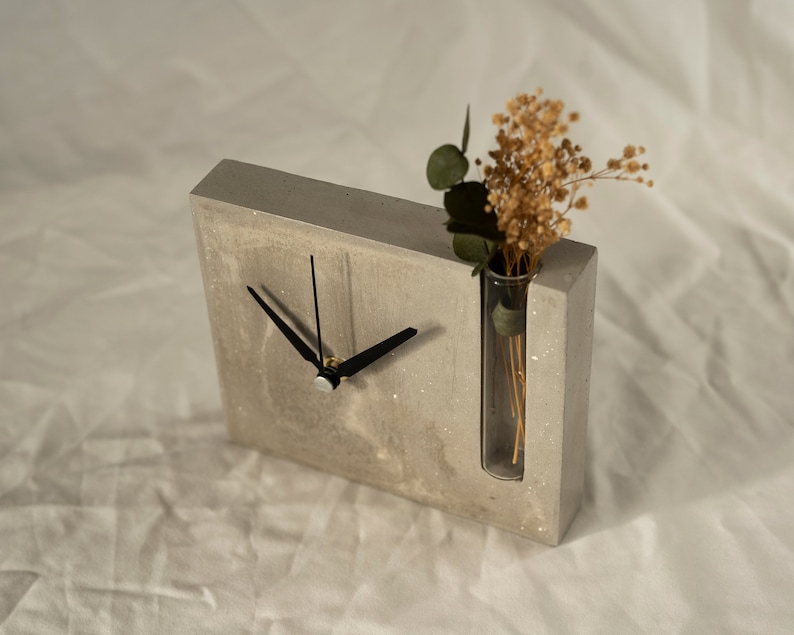 May include: A gray concrete clock with black hands and a glass tube vase for dried flowers. The vase is built into the clock face.