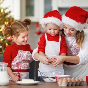 Pu&ograve; includere: Una famiglia con cappelli di Babbo Natale rossi e bianchi cuoce biscotti di Natale in cucina. Una bambina con un grembiule rosso e bianco guarda una donna rompere un uovo in una ciotola. Un bambino con un grembiule rosso e bianco tiene l'uovo.