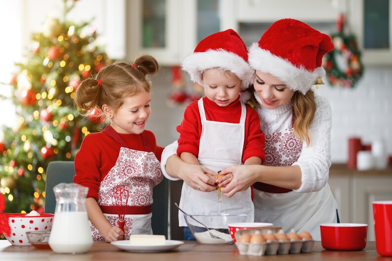 Pu&ograve; includere: Una famiglia con cappelli di Babbo Natale rossi e bianchi cuoce biscotti di Natale in cucina. Una bambina con un grembiule rosso e bianco guarda una donna rompere un uovo in una ciotola. Un bambino con un grembiule rosso e bianco tiene l'uovo.