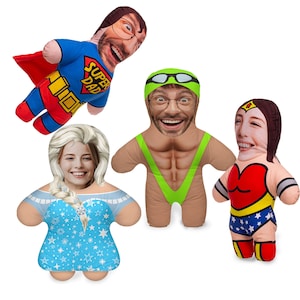 Peut inclure: Quatre peluches avec des visages de personnes. Les peluches sont habillées en costumes. L'une est un super-héros avec un costume rouge et bleu, l'autre est une femme en costume de Wonder Woman rouge et bleu, l'une est un homme en maillot de bain vert et l'autre est une femme en robe bleue avec un motif de flocons de neige blancs.