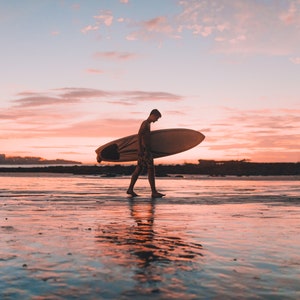 Puede incluir: Una persona camina por una playa al atardecer llevando una tabla de surf. El cielo es de un rosa y naranja vibrantes.