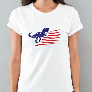 以下が含まれることがあります： 赤白青のアメリカ国旗のデザインが施された白いTシャツ。ティラノサウルス・レックスのシルエットが描かれている。