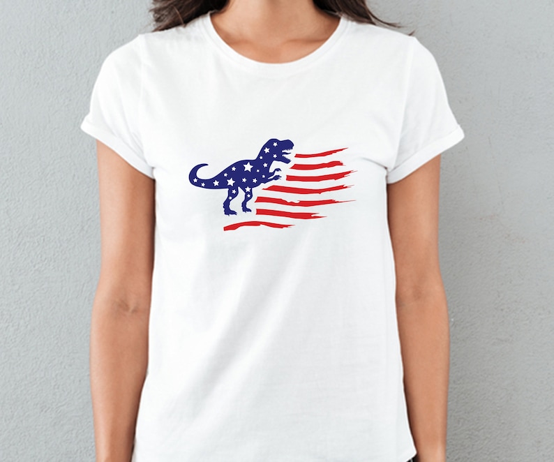 以下が含まれることがあります： 赤白青のアメリカ国旗のデザインが施された白いTシャツ。ティラノサウルス・レックスのシルエットが描かれている。