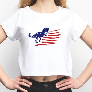 以下が含まれることがあります： 青と赤のアメリカ国旗のデザインが施された白いクロップドTシャツ。恐竜のシルエットが描かれています。