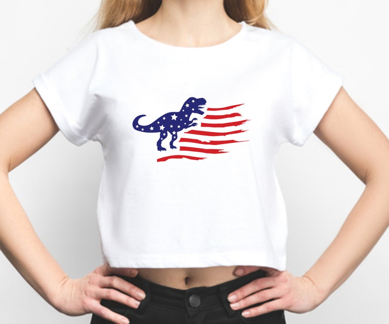 以下が含まれることがあります： 青と赤のアメリカ国旗のデザインが施された白いクロップドTシャツ。恐竜のシルエットが描かれています。