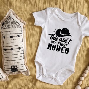 May include: White baby onesie with black text that reads "This ain't my first rodeo" with a black cowboy hat graphic above the text.