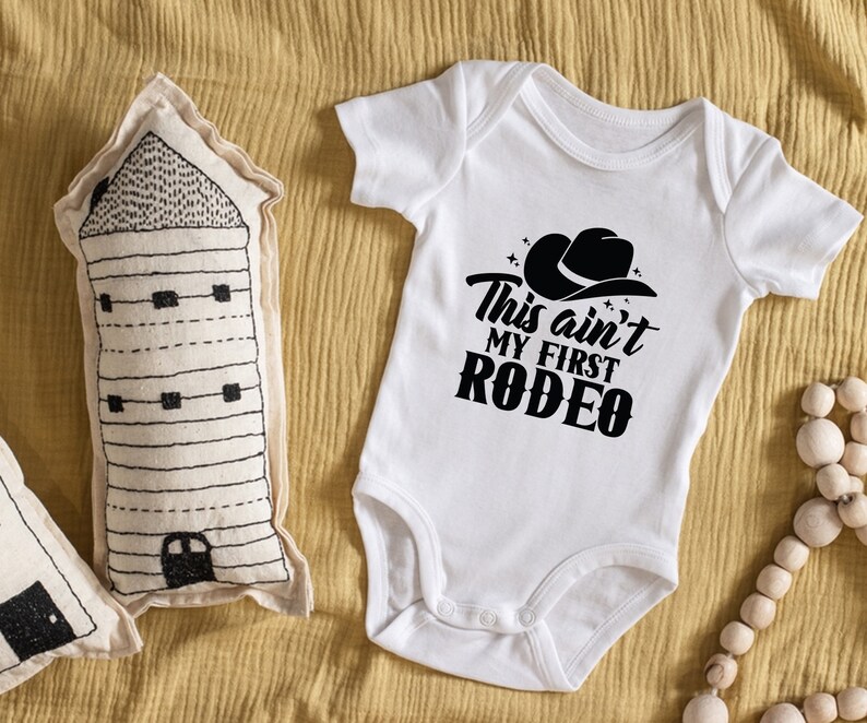 May include: White baby onesie with black text that reads "This ain't my first rodeo" with a black cowboy hat graphic above the text.