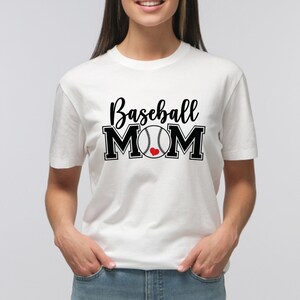 May include: White t-shirt with the words "Baseball MOM" in black script and block letters. A baseball graphic with a red heart replaces the "O" in MOM. The shirt is short-sleeved and has a crew neck.