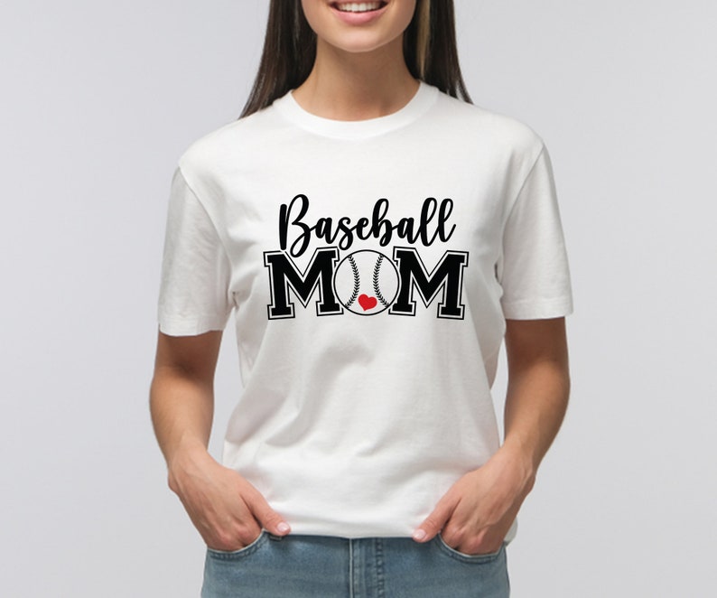 May include: White t-shirt with the words "Baseball MOM" in black script and block letters. A baseball graphic with a red heart replaces the "O" in MOM. The shirt is short-sleeved and has a crew neck.