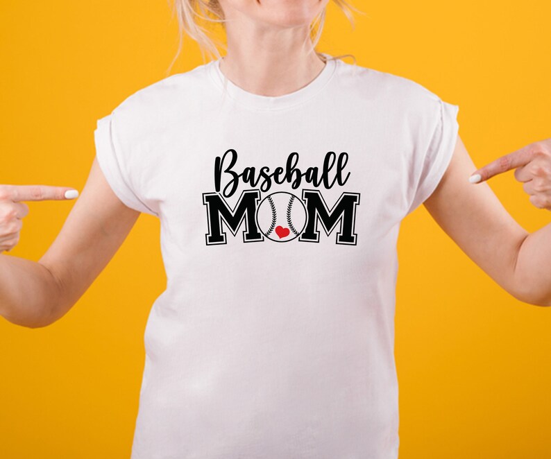 May include: White t-shirt with the words "Baseball MOM" in black script and block letters. A baseball with a red heart is in the center of the word "MOM". The shirt is on a person with a yellow background.
