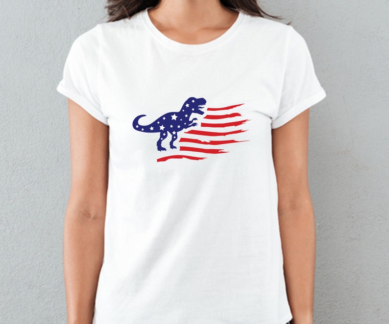 以下が含まれることがあります： 赤白青のアメリカ国旗のデザインが施された白いTシャツ。ティラノサウルス・レックスのシルエットが描かれている。