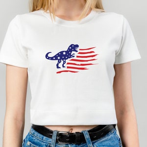 以下が含まれることがあります： 赤、白、青のアメリカ国旗のデザインが施された白いクロップドTシャツ。青い恐竜のシルエットが特徴です。
