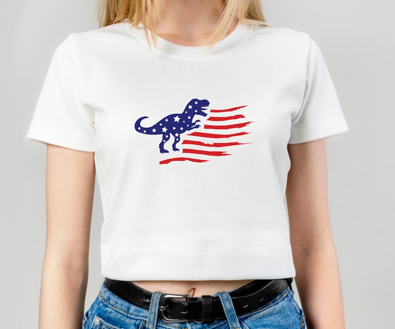 以下が含まれることがあります： 赤、白、青のアメリカ国旗のデザインが施された白いクロップドTシャツ。青い恐竜のシルエットが特徴です。