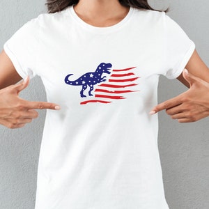 以下が含まれることがあります： 赤、白、青のアメリカ国旗のデザインが施された白いTシャツ。青い恐竜のシルエットが描かれている。