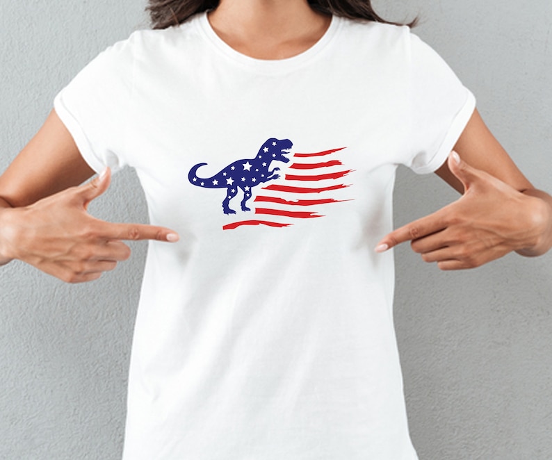 以下が含まれることがあります： 赤、白、青のアメリカ国旗のデザインが施された白いTシャツ。青い恐竜のシルエットが描かれている。