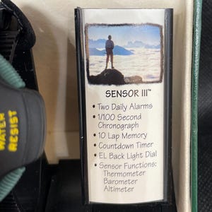 K&ouml;nnte beinhalten: Eine schwarz-gr&uuml;ne Uhr mit dem Text "WATER RESIST" an der Seite. Die Uhr befindet sich in einer gr&uuml;nen Box mit einer Karte mit der Aufschrift "SENSOR III" und listet Funktionen wie zwei t&auml;gliche Alarme, einen Chronographen und Sensorfunktionen auf.