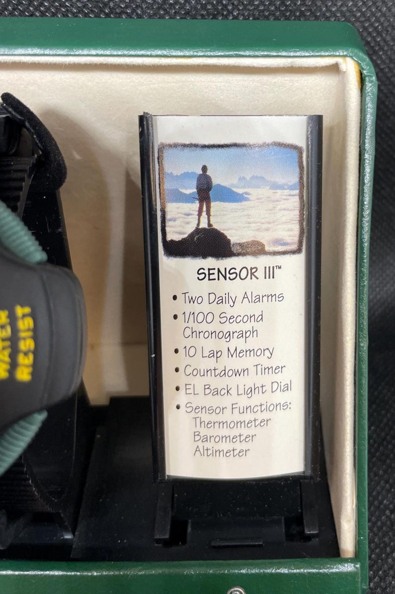 K&ouml;nnte beinhalten: Eine schwarz-gr&uuml;ne Uhr mit dem Text "WATER RESIST" an der Seite. Die Uhr befindet sich in einer gr&uuml;nen Box mit einer Karte mit der Aufschrift "SENSOR III" und listet Funktionen wie zwei t&auml;gliche Alarme, einen Chronographen und Sensorfunktionen auf.