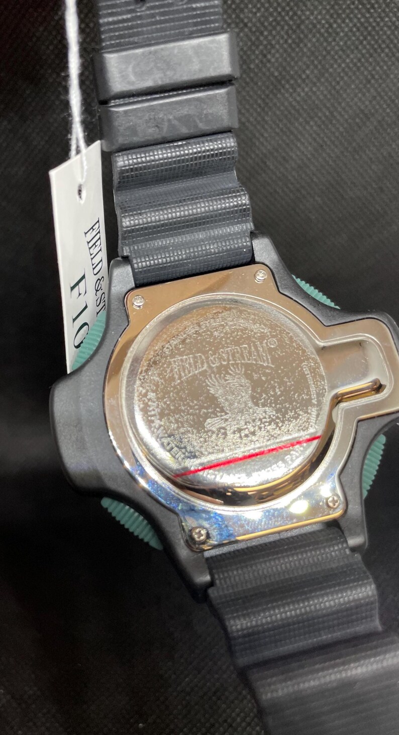 K&ouml;nnte beinhalten: Schwarze Uhr mit schwarzem Armband und silbernem R&uuml;cken. Die Uhr hat einen gr&uuml;nen Akzent an der Seite. Auf der R&uuml;ckseite der Uhr stehen die Worte "FIELD & STREAM" und ein Adlerbild. Ein wei&szlig;es Etikett ist angebracht.