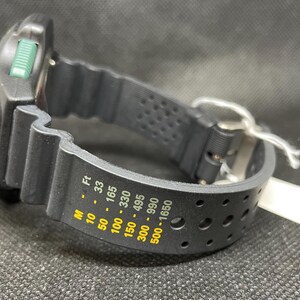 K&ouml;nnte beinhalten: Schwarze und silberne Taucheruhr mit schwarzem Kautschukarmband. Das Zifferblatt der Uhr hat gr&uuml;ne Akzente und eine Digitalanzeige. Das Armband hat Tiefenmarkierungen in Fu&szlig; und Meter. Ein wei&szlig;es Etikett ist am Armband befestigt.
