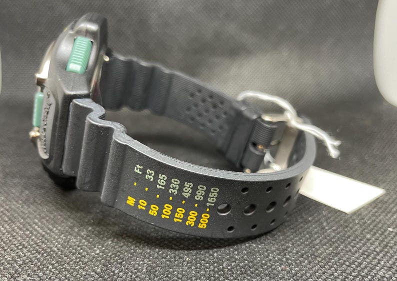 K&ouml;nnte beinhalten: Schwarze und silberne Taucheruhr mit schwarzem Kautschukarmband. Das Zifferblatt der Uhr hat gr&uuml;ne Akzente und eine Digitalanzeige. Das Armband hat Tiefenmarkierungen in Fu&szlig; und Meter. Ein wei&szlig;es Etikett ist am Armband befestigt.