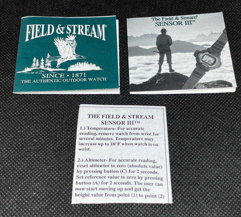 K&ouml;nnte beinhalten: Drei bedruckte Karten. Eine ist t&uuml;rkis und tr&auml;gt die Aufschrift "FIELD & STREAM" und "SINCE 1871 THE AUTHENTIC OUTDOOR WATCH". Eine andere zeigt eine Uhr und eine Person auf einem Berg. Die dritte Karte enth&auml;lt Anweisungen f&uuml;r die Uhr "SENSOR III".
