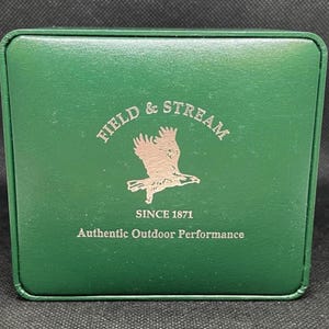 K&ouml;nnte beinhalten: Eine gr&uuml;ne Box mit silbernem Schriftzug "FIELD & STREAM" und einem Adlerlogo. Der Text "SINCE 1871" und "Authentic Outdoor Performance" sind ebenfalls abgebildet.