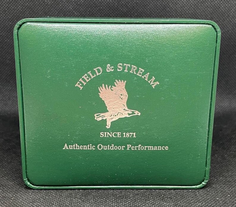K&ouml;nnte beinhalten: Eine gr&uuml;ne Box mit silbernem Schriftzug "FIELD & STREAM" und einem Adlerlogo. Der Text "SINCE 1871" und "Authentic Outdoor Performance" sind ebenfalls abgebildet.