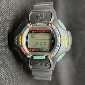 K&ouml;nnte beinhalten: Schwarze Digitaluhr mit silberner L&uuml;nette und schwarzem Armband. Das Zifferblatt zeigt die Uhrzeit und das Datum an. Die Uhr hat Kn&ouml;pfe mit der Aufschrift "Sensor", "Mode", "Adj", "St/Stp", "Alti-Baro" und "Water Resist". Der Markenname "Field & Stream" ist sichtbar.