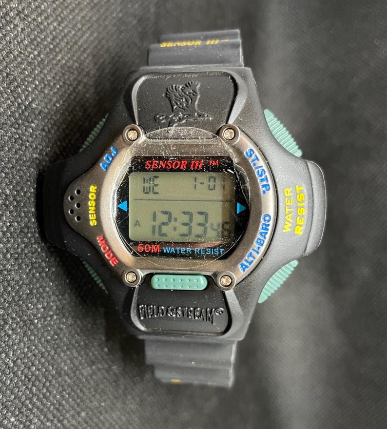 K&ouml;nnte beinhalten: Schwarze Digitaluhr mit silberner L&uuml;nette und schwarzem Armband. Das Zifferblatt zeigt die Uhrzeit und das Datum an. Die Uhr hat Kn&ouml;pfe mit der Aufschrift "Sensor", "Mode", "Adj", "St/Stp", "Alti-Baro" und "Water Resist". Der Markenname "Field & Stream" ist sichtbar.