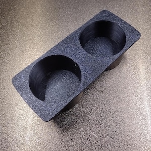 Op de afbeelding: Zwarte plastic bekerhouder met twee ronde openingen. De houder heeft een getextureerd oppervlak.