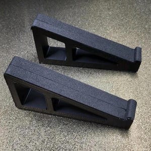 May include: Two black, angled car ramps with a textured surface. Each ramp has a rectangular opening and a curved lip at the end. The ramps are designed to elevate a vehicle for maintenance or repairs.
