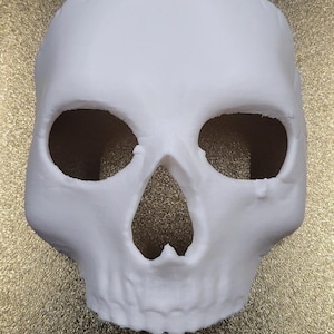 Puede incluir: Una máscara de calavera blanca con cuencas de ojos grandes y una cavidad nasal prominente. La máscara está hecha de un material liso y mate y está diseñada para adaptarse a la cara. La máscara de calavera es un accesorio de Halloween o disfraz.