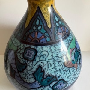 Peut inclure: Un vase en céramique décoratif avec un corps bulbeux et un col étroit. Le vase présente un bord jaune et un motif détaillé de motifs floraux turquoise, bleus et violets. Le fond est de couleur turquoise clair.