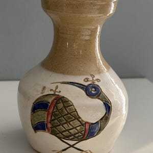 May include: A ceramic vase with a flared rim and a speckled tan neck. The body is off-white and features a hand-painted graphic of a bird with a long curved beak, blue and green patterned wings, and red accents, standing on two thin dark lines.