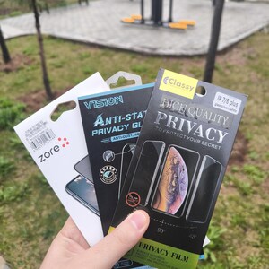 Puede incluir: Una mano sostiene tres paquetes de protectores de pantalla para teléfonos. Los paquetes tienen el siguiente texto: "zore", "VISION Anti-Static Privacy GL" y "Classy High Quality Privacy". El fondo es un parque.