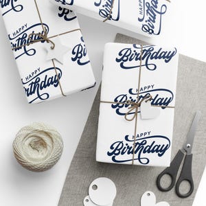 May include: White gift-wrapped boxes with navy blue "Happy Birthday" text, tied with twine and decorative tags. A ball of twine, scissors, and gift tags are also present, suggesting a celebratory theme.