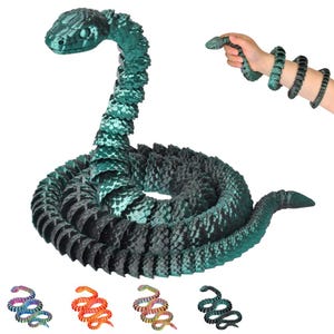 Op de afbeelding: Een groot, geleed slangen speelgoed in tinten teal en zwart, opgerold op een witte achtergrond. De kop van de slang is omhoog, en een kleinere versie wordt in een hand gehouden. Extra kleinere slangen speelgoed in verschillende kleuren worden hieronder getoond.