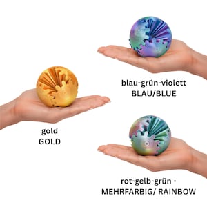 Op de afbeelding: Drie kleurrijke fidget-speeltjes in de vorm van tandwielen worden getoond. Eén is goudkleurig, een andere is blauw-groen-violet en de derde is rood-geel-groen. De tekst "gold", "blau-grün-violett BLAU/BLUE" en "rot-gelb-grün - MEHRFARBIG/ RAINBOW" is ook zichtbaar.