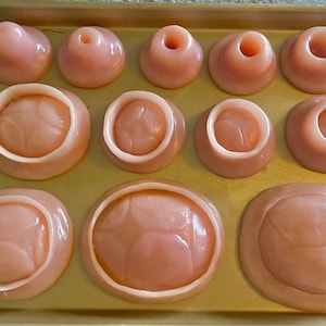 Puede incluir: Una colección de objetos rosados y redondeados de diversas formas y tamaños, dispuestos sobre una bandeja dorada. Algunos tienen un agujero central, otros son sólidos o tienen un borde elevado. Los objetos parecen estar hechos de un material liso, posiblemente comestible.