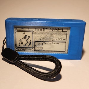 May include: A blue handheld electronic device with a black and white screen displaying a pixelated image of a creature. The device has a black wrist strap and the text "ResCuegotchi" is visible on the screen.