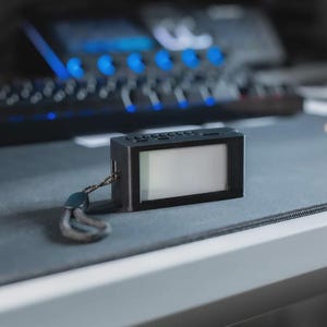 Puede incluir: Un pequeño dispositivo rectangular negro con pantalla y correa de muñeca. El dispositivo está sobre una superficie gris, con un teclado y luces azules en el fondo. La pantalla está en blanco.