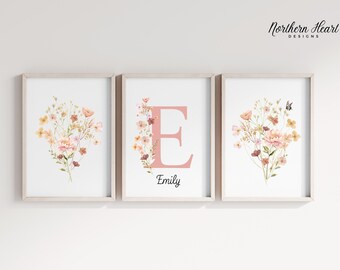 May include: Three framed prints with a floral design. The center print features a large pink letter "E" with the name "Emily" below it. The prints are on a white wall.