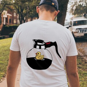 May include: White t-shirt with a cartoon coffee pot design and a yellow rubber duck inside. The coffee pot has a black handle and a red accent. The person is wearing a blue baseball cap.