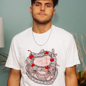 May include: White t-shirt featuring a pink and white cake design with red cherries and the text "Eat The Rich." The graphic tee is worn by a person with short dark hair and a silver chain necklace.