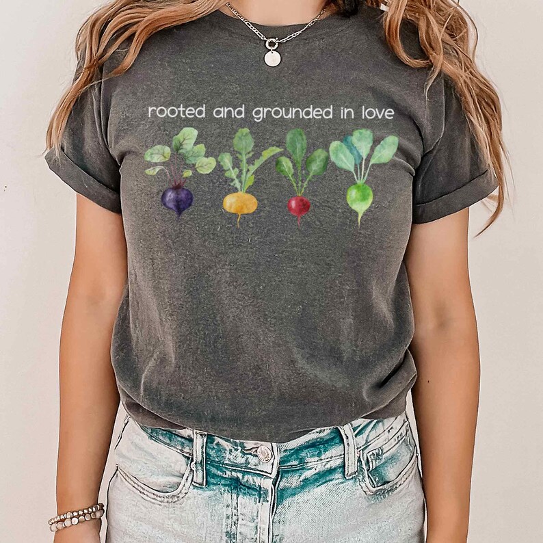 May include: A dark gray t-shirt with a white graphic of five colorful root vegetables and the text "rooted and grounded in love".