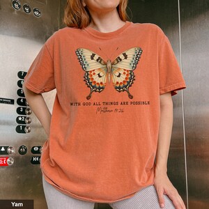 Puede incluir: Una camiseta naranja quemada con un gr&aacute;fico de mariposa y el texto "Con Dios todo es posible Mateo 19:26".