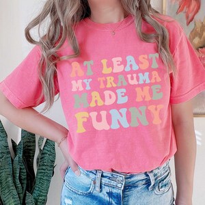 May include: Pink t-shirt with the text "At Least My Trauma Made Me Funny" in rainbow colours.