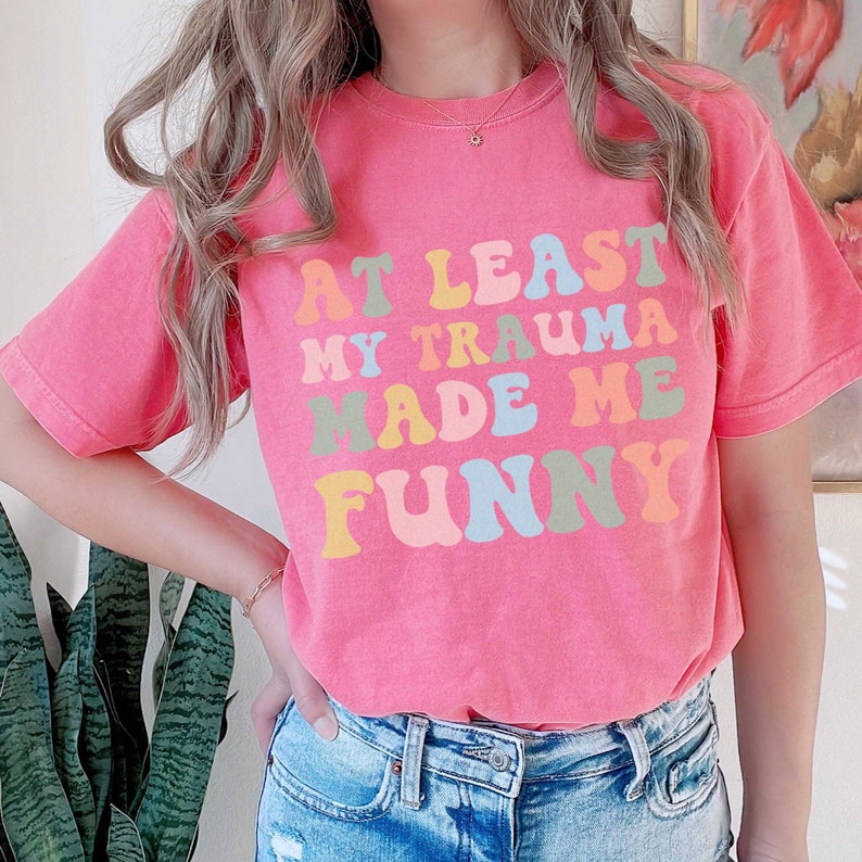 May include: Pink t-shirt with the text "At Least My Trauma Made Me Funny" in rainbow colours.