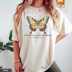 Puede incluir: Una camiseta de color crema con un gr&aacute;fico de mariposa colorida y el texto "Con Dios todo es posible Mateo 19:26".